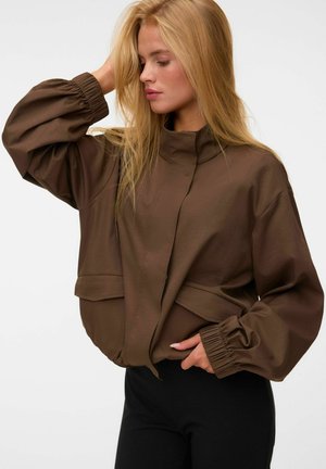 Femme blonde portant une veste marron ample avec des poignets élastiques, posant avec une main sur la tête et l'autre dans la poche.
