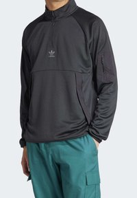 Man som bär en svart Adidas tröja med kvartslång dragkedja och ficka med dragkedja på ärmen samt teal-färgade cargobyxor, med händerna i fickorna, vänd framåt.