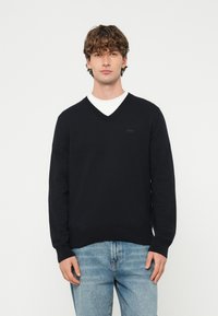 Młody mężczyzna o kręconych włosach, ubrany w czarny sweter z dekoltem w serek na białą koszulę i jasnoniebieskie dżinsy, stoi na jednolitym tle.