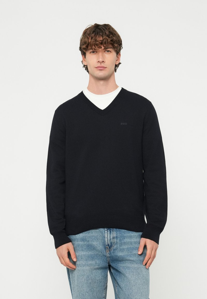 Młody mężczyzna o kręconych włosach, ubrany w czarny sweter z dekoltem w serek na białą koszulę i jasnoniebieskie dżinsy, stoi na jednolitym tle.