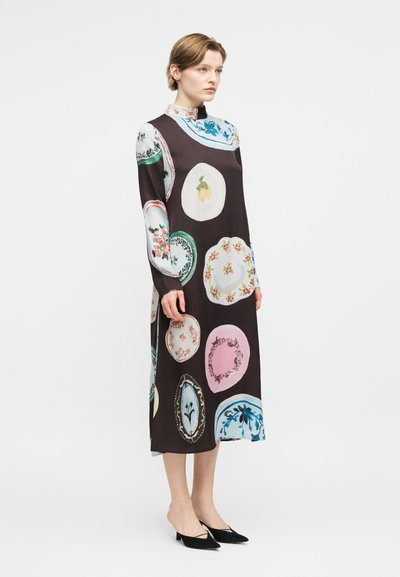 Γυναίκα που στέκεται με μακρύ μαύρο φόρεμα διακοσμημένο με πολύχρωμα μοτίβα από πιάτα, φοράει μαύρα μυτερά παπούτσια με χαμηλό τακούνι σε λευκό φόντο.