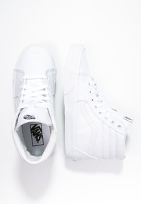 High-top white sneakers made of canvas, featuring a textured side, round toe, flat sole, laces, and logo on the tongue.