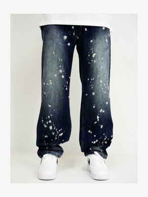 Blaue Denim-Jeans mit lockerer Passform, verziert mit weißen Farbspritzern. Kombiniert mit weißen Turnschuhen. Details umfassen Standardtaschen und Nähte.
