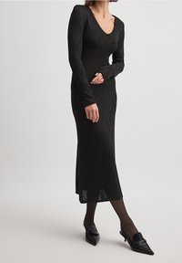 Femme portant une longue robe en maille texturée noire à manches longues, des collants noirs transparents et des mocassins à talons aigus noirs.