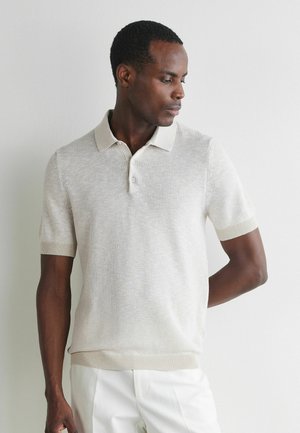 Mann trägt beige kurzärmliges Strick-Poloshirt und weiße Hose, steht vor einer schlichten hellen Wand und schaut nach links.