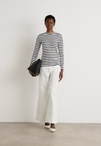 Camicia a maniche lunghe a righe in bianco e nero, abbinata a pantaloni bianchi a gamba larga. La modella tiene in mano una pochette nera e indossa tacchi bianchi.
