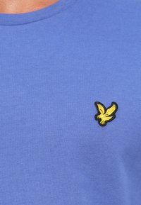 T-shirt en coton violet à manches courtes, avec un logo d'aigle brodé jaune à bord noir sur la zone de la poitrine gauche.