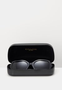 Des lunettes de soleil Coach noires dans un étui en cuir noir. Les lunettes ont un design audacieux avec des verres foncés et une finition lisse. Logo doré à l'intérieur de l'étui.
