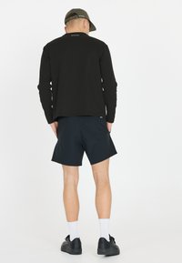 Schwarzes Langarmshirt und marineblaue Shorts, mit einem dezenten Logo auf der Rückseite des Shirts. Das Model trägt schwarze Schuhe und weiße Socken.