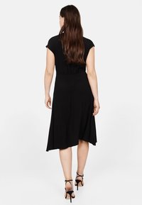 Schwarzes Kleid mit kurzen Ärmeln, tailliertem Schnitt und ausgestelltem Rock. Glattes Material mit knielangem Schnitt, kombiniert mit schwarz geschnürten High Heels.