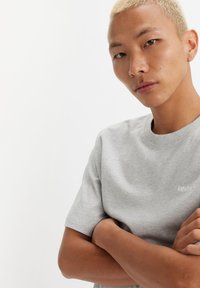 Pelēkā kokvilnas T-kreklā īsas piedurknes, apaļa kakla izgriezums un neliels balts Levi's logo uz krūtīm. Modelis ar rokām saliktām krūtīs.