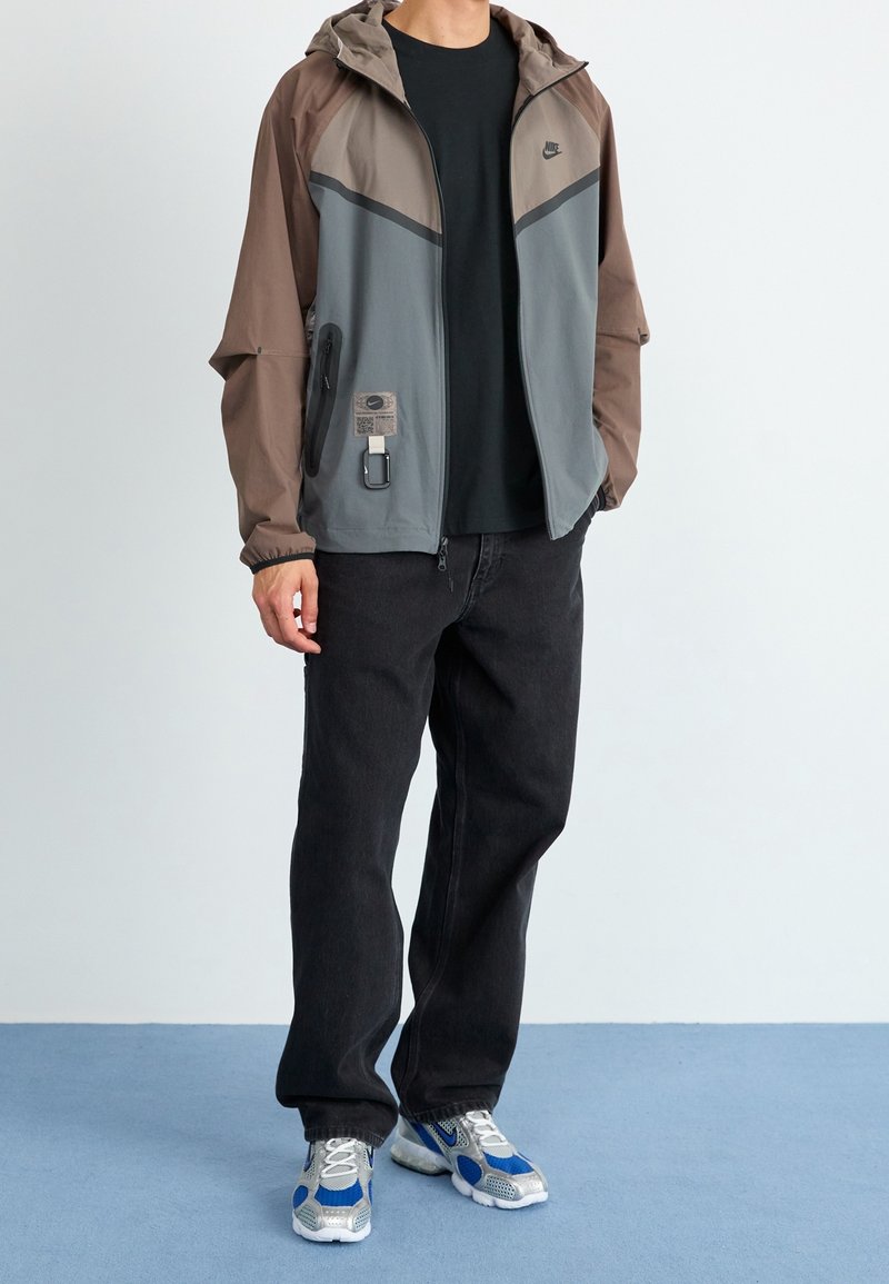Personne portant une veste zippée Nike marron et grise, un t-shirt noir, un jean noir et des baskets argentées et bleues, debout sur un sol bleu.