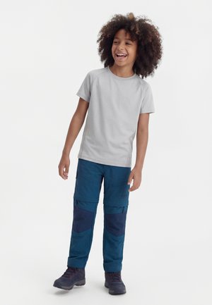 Dieťa s kučeravými vlasmi sa usmieva, má na sebe svetlosivé tričko s krátkym rukávom, modré cargo nohavice s náplasťami na kolenách a tmavé turistické topánky, stojí na bielom pozadí.