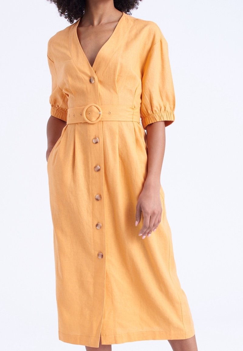 Femme portant une robe jaune mi-longue avec des boutons sur le devant, des manches courtes bouffantes et une ceinture assortie avec une boucle ronde.