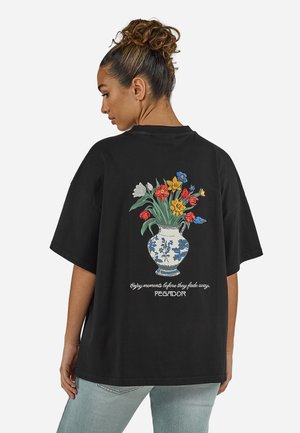 Woman with curly hair in a bun wearing a black oversized t-shirt with a colorful floral vase and text on the back.