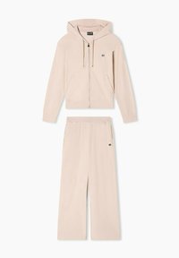 Sudadera con cremallera de color beige claro, con capucha ajustable mediante cordón y logo en el pecho, combinada con pantalones de pierna ancha a juego que tienen bolsillos laterales.