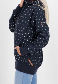 Kvinna som bär en marinblå zip-up jacka med vitt fågelmönster och ribbade manschetter, tillsammans med ljusblå jeans, med handen i jackfickan.