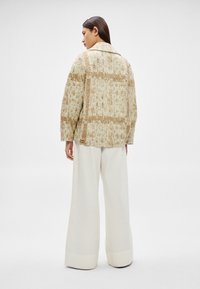 Sieviete stāv, valkājot bēša krāsas teksturētu žaketi ar rūtainu rakstu un platas balta auduma bikses, skatu pagriezusi prom uz koptu fona.