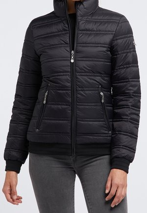 Frau trägt eine schwarze gesteppte Jacke mit Frontreißverschluss und Reißverschlusstaschen, kombiniert mit grauer Hose, vor einem einfarbigen Hintergrund.