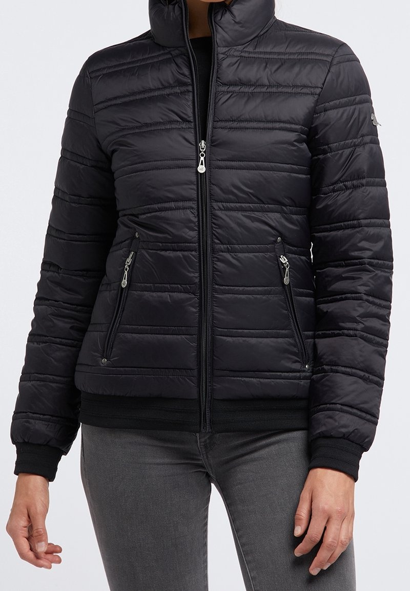 Femme portant une veste noire matelassée avec fermeture éclair avant et poches zippées, associée à un pantalon gris, sur un fond uni.