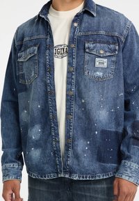 Donkerblauwe denimjas met lichtere spetterpatronen, knoopsluiting, twee borstzakken en een patchdetail aan de rechterkant.