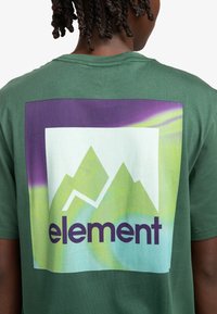 Camiseta de algodón verde con un gran gráfico en la parte posterior que presenta montañas estilizadas en verde claro y púrpura, y la palabra "element" en púrpura oscuro.