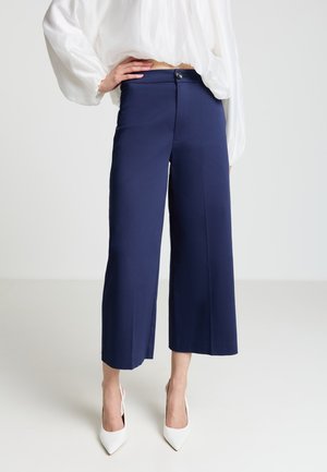 Culottes blu navy realizzate in tessuto morbido, con chiusura a bottoni e gambe ampie che arrivano a metà polpaccio. Abbinate a décolleté bianchi con punta appuntita.