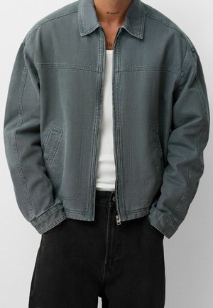 Homme portant une veste grise zippée sur un débardeur blanc avec un pantalon noir, les mains dans les poches de la veste.