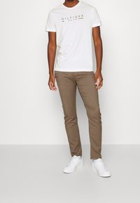 Muž v bílém tričku Hilfiger New York, hnědých úzkých kalhotách a bílých teniskách stojící na jednobarevném světlém pozadí.