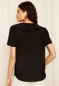 Top negru dintr-un material mesh texturat, cu mâneci scurte tip balon și un detaliu cu volane la decolteu, purtat cu pantaloni de o culoare deschisă.