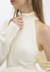 Top bianco a costine con collo alto e chiusura con zip, con una spalla scoperta e maniche lunghe. Texture morbida con un leggero lucido.