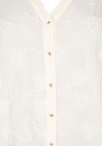 Blusa bianca con tessuto strutturato, decorata con ricami floreali, chiusura con bottoni e bottoni dorati lungo la parte anteriore. Design a scollo a V.