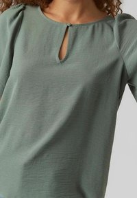 Blusa verde a maniche corte con scollatura a forma di chiave. Tessuto leggermente testurizzato, maniche a sbuffo e dettaglio decorativo con bottone al collo.