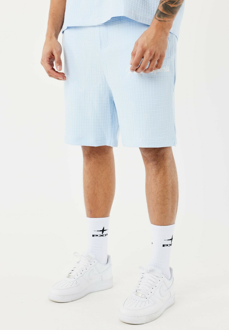 Pantaloni corti azzurro chiaro in tessuto testurizzato, con tasche e dettagli del logo discreti. Abbinati a sneakers bianche e calze a fantasia.