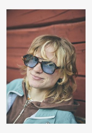 Large, square sunglasses with blue tinted lenses and a tortoiseshell frame. Worn with a green zip-up jacket over a brown top.