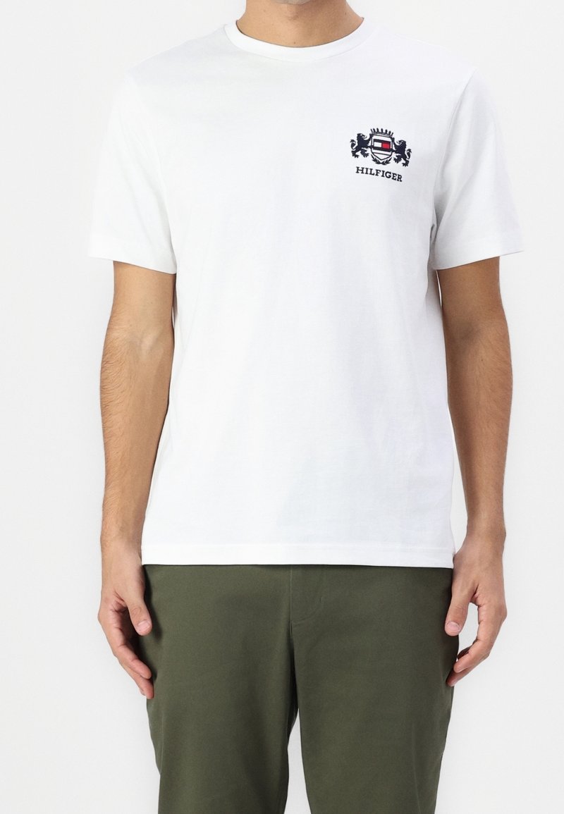 Homme portant un t-shirt blanc à manches courtes avec un petit logo de blason noir et rouge et un texte sur le côté gauche de la poitrine, associé à un pantalon vert foncé.