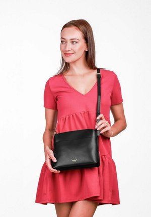 Černá kožená kabelka přes rameno s hladkým povrchem, vybavená horním zipem, stříbrnými doplňky a nastavitelným popruhem. Modelka má na sobě růžové šaty.