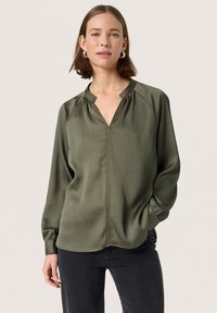 Blusa verde oliva con una texture ondulata, scollatura a V e maniche lunghe. Presenta dettagli arricciati sulle spalle e una vestibilità ampia.