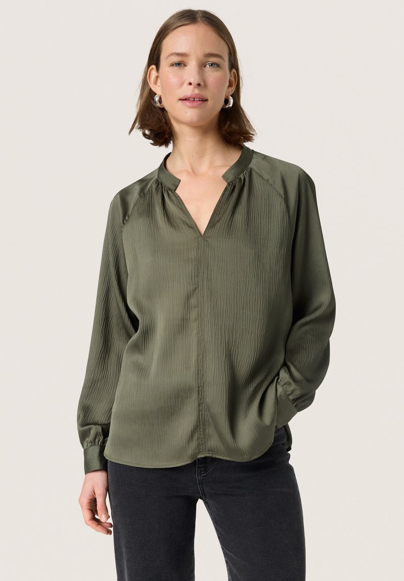 Blusa verde oliva con una texture ondulata, scollatura a V e maniche lunghe. Presenta dettagli arricciati sulle spalle e una vestibilità ampia.