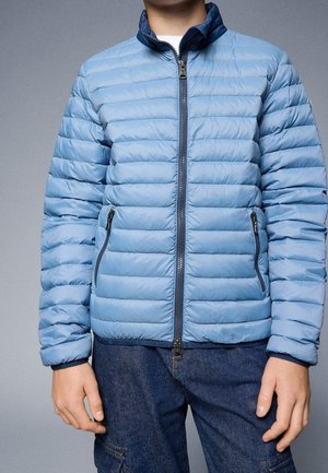 Personne portant une veste matelassée bleu clair avec une fermeture éclair frontale et des poches zippées, associée à un jean bleu foncé.