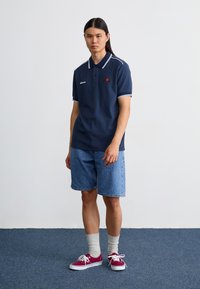 Námornícky polo tričko s bielym lemovaním, vyšívané logo, v páre s denimovými šortkami. Model má na sebe marónové tenisky a šedé ponožky. Jednoduché pozadie.