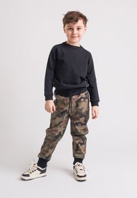 Młody chłopiec stoi w czarnej bluzie, spodniatch w kamuflaż oraz czarno-białych trampkach na jednolitym jasnym tle.