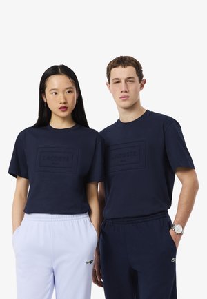Due modelli indossano t-shirt Lacoste blu navy; uno con pantaloni della tuta azzurri e l'altro con pantaloni della tuta blu navy abbinati, entrambi con le mani nelle tasche.