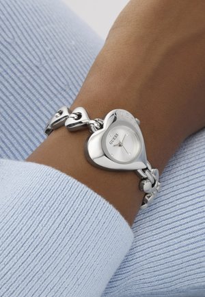 Zilverkleurig horloge in de vorm van een hart met een schakelkettinkje, voorzien van een witte wijzerplaat en vier kleine kristallen op de wijzerplaat, geplaatst op een beige pols.