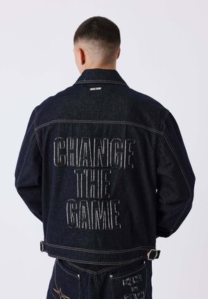 Personne aux cheveux courts portant une veste en jean foncée et un jean, le dos de la veste présente un grand texte brodé "CHANGE THE GAME" en fil blanc.