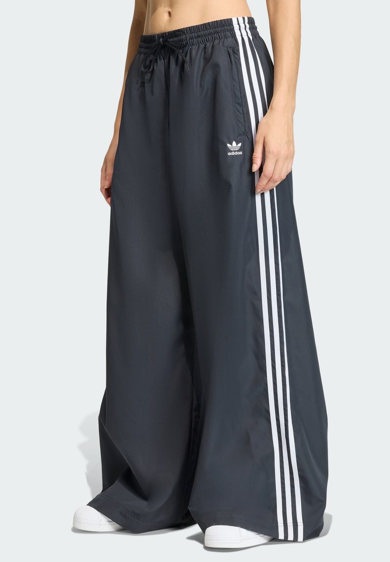 Pantalones negros de pierna ancha hechos de tela ligera con rayas blancas de Adidas en los lados y una cinturilla elástica. Se llevan con zapatos blancos.