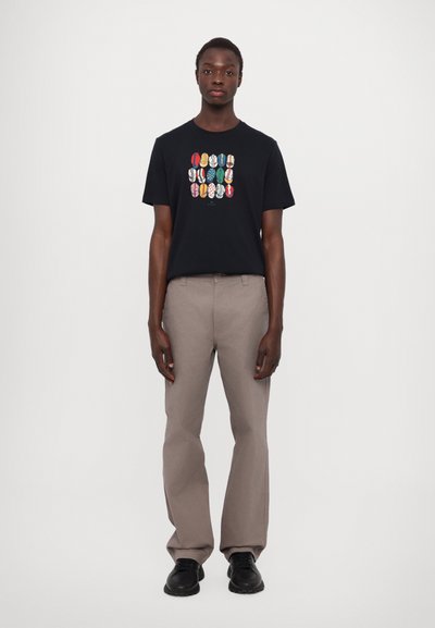 T-shirt en coton noir avec un design graphique coloré au centre. Associé à un pantalon droit gris clair et des chaussures noires.