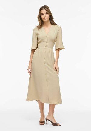 Kvinde står iført en beige midi-kjole med albuelange ærmer og knapper foran, kombineret med brune hælede sandaler med åben tå.