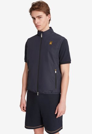 Junger Mann trägt eine dunkelblaue ärmellose Weste mit Reißverschluss über einem kurzärmligen Hemd und schwarzen Shorts, steht vor einem weißen Hintergrund.