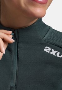 Mørkegrøn sports-top med høj krave, delvis lynlås, strukturerede skulderdetaljer og hvid "2XU"-logo på brystet.